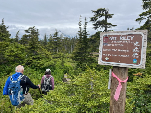 Two hikers on a trail near a sign for Mt. Riley, surrounded by lush greenery and trees.