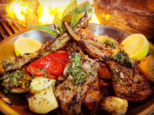 A sizzling platter of grilled lamb chops with herbs, roasted vegetables, and lemon, set against a backdrop of flames.
