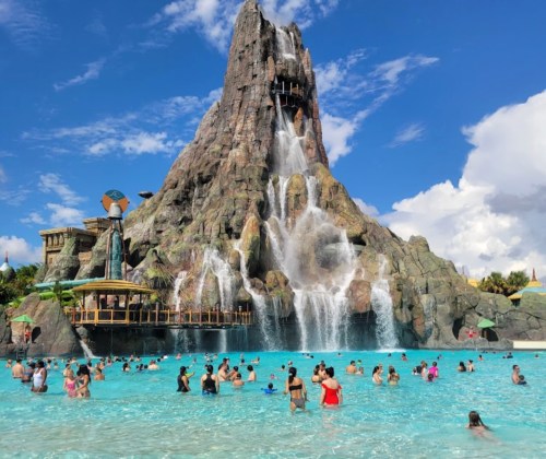 A large volcanic structure with waterfalls towers over a crowded water park pool on a sunny day.
