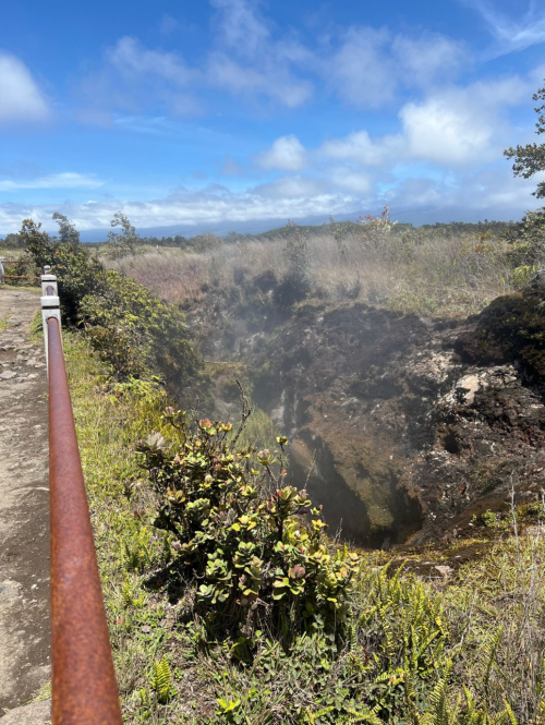 A volcanic landscape with steam rising from a crater, surrounded by lush vegetation and a clear blue sky.