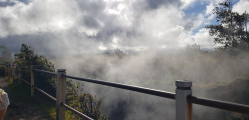 A misty landscape with a railing, surrounded by clouds and trees under a partly cloudy sky.