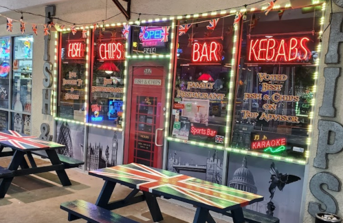 A brightly lit restaurant exterior featuring neon signs for fish and chips, kebabs, and karaoke, with Union Jack decor.