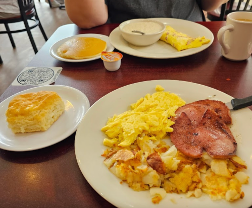 A breakfast spread featuring scrambled eggs, ham, hash browns, a biscuit, and a pancake on a table.