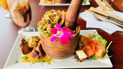 A wooden bowl filled with shrimp and sauce, garnished with an orchid, alongside salad and grilled meat on a table.