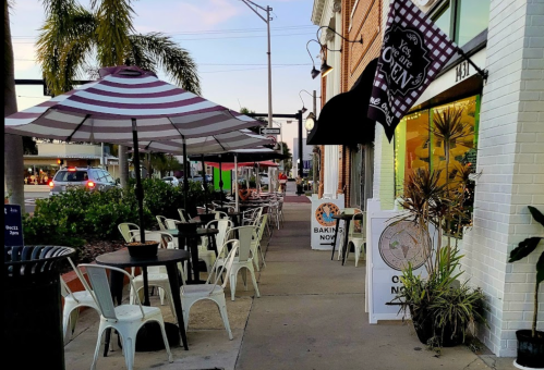 Outdoor café scene with striped umbrellas, empty tables, and palm trees along a sidewalk at dusk.