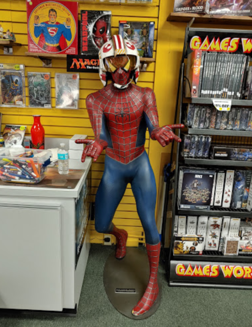 A life-sized Spider-Man statue wearing a helmet, displayed in a comic shop surrounded by games and collectibles.