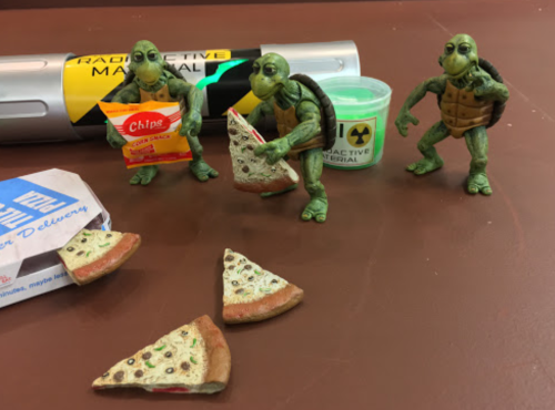 Three turtle figurines holding pizza slices and a snack bag, with a container labeled "Radioactive Material" nearby.