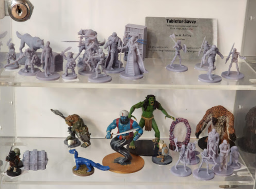 A display of various unpainted tabletop miniatures, including fantasy and sci-fi characters, on a clear shelf.