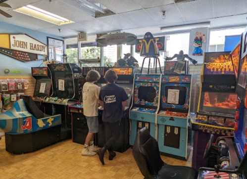 Two people stand at vintage arcade machines in a colorful game shop, with various decorations and posters in the background.