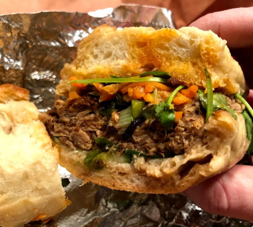 A close-up of a sandwich filled with shredded meat, fresh herbs, and vegetables, held in a hand.