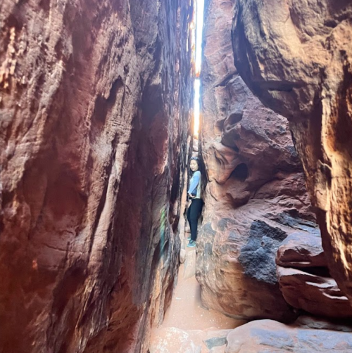 A person stands between two towering rock formations in a narrow canyon, illuminated by sunlight above.