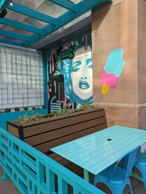 A colorful outdoor seating area with a mural of a woman, a payphone, and a large ice cream pop decoration.