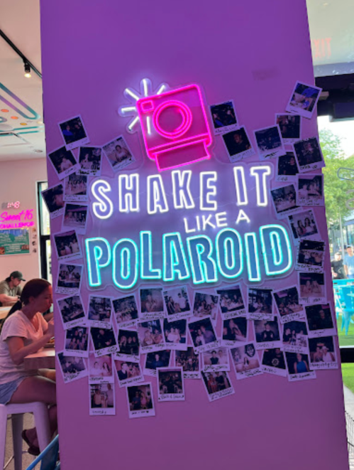 Neon sign reading "Shake it like a Polaroid" surrounded by Polaroid photos on a colorful wall.