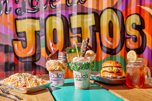 Colorful milkshakes and a burger on a table, with a vibrant mural reading "JOTOS" in the background.