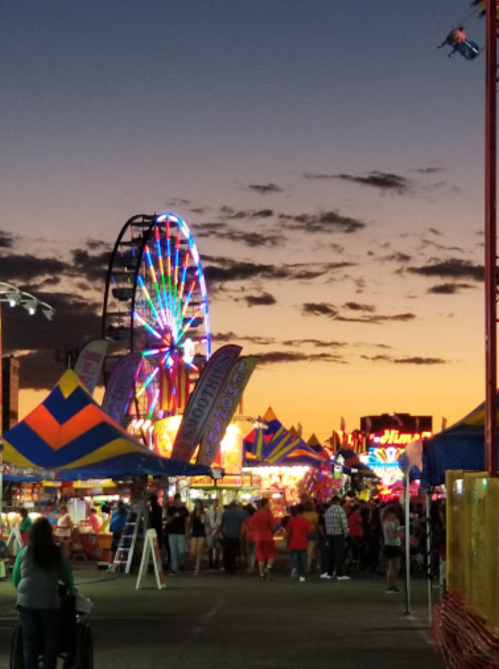 A vibrant carnival scene at dusk, featuring a Ferris wheel and colorful tents under a sunset sky.