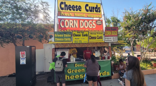 A food stand featuring cheese curds and corn dogs, with customers ordering at the counter. Bright signage displays menu items.