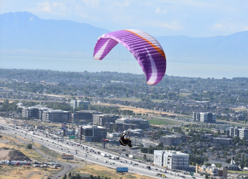 A paraglider with a purple canopy soars above a cityscape and highway, with mountains in the background.