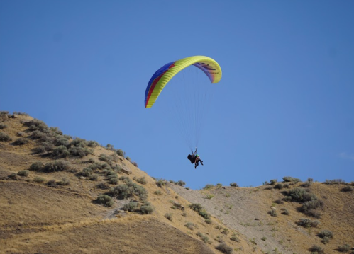 A person paragliding over a dry, hilly landscape under a clear blue sky.