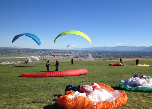 Paragliders launch from a grassy hill, colorful canopies against a clear blue sky, with a scenic view below.