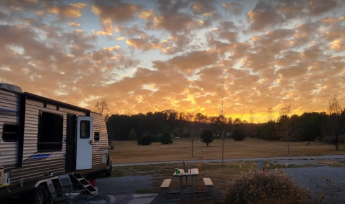 A scenic sunset with colorful clouds over a grassy field, featuring an RV and a picnic table in the foreground.