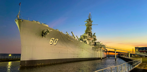 A battleship docked at sunset, with a clear sky and water reflecting the warm colors of the evening.