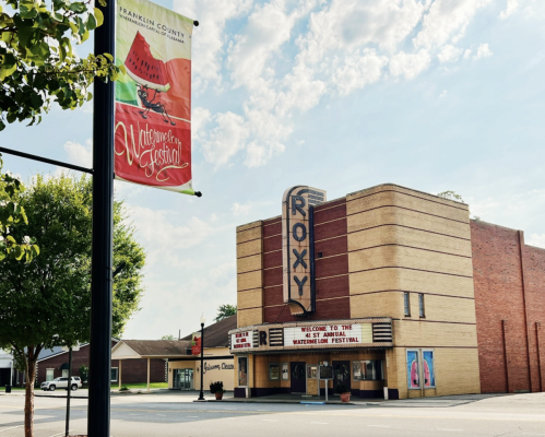 A vintage theater building with a "Roxy" sign, surrounded by trees and a banner for a watermelon festival.