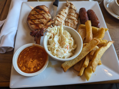 A plate with grilled chicken, sausages, coleslaw, fries, and a small bowl of sauce.