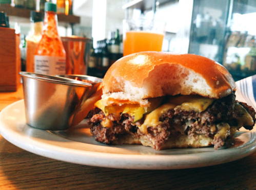A close-up of a cheeseburger with a side of dipping sauce on a plate, set in a casual dining environment.