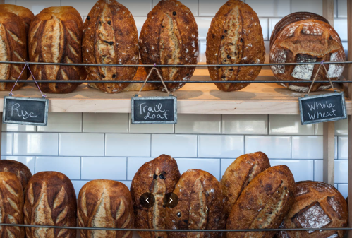 A display of various artisanal bread loaves, including rye, trail loaf, and whole wheat, on wooden shelves.