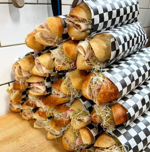 A stack of sandwiches wrapped in black and white checkered paper, filled with meats and sprouts.