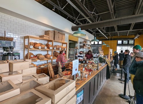 A busy bakery with a variety of breads and pastries displayed, customers in line, and staff serving at the counter.