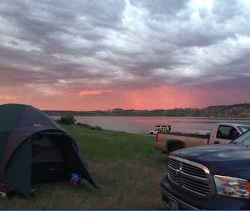 A campsite by a lake at sunset, featuring a tent, a truck, and dramatic clouds in the sky.