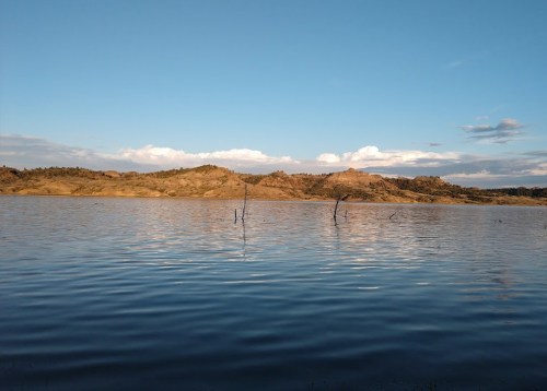 A serene lake reflects the sky, surrounded by rolling hills and sparse trees under a clear blue sky.