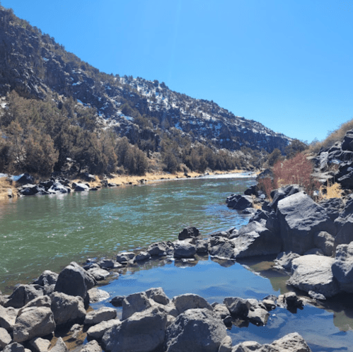 A serene river flows between rocky banks, surrounded by trees and mountains under a clear blue sky.