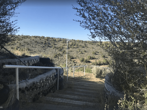 A pathway with wooden steps leads down through shrubs to a hilly landscape under a clear blue sky.