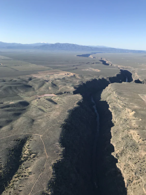 Aerial view of a deep canyon surrounded by vast, flat terrain and distant mountains under a clear blue sky.
