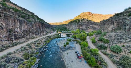 A scenic view of a river winding through rocky terrain, with a bridge and trees along the banks under a clear blue sky.