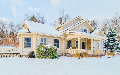 A cozy house with a porch, surrounded by snow-covered ground and trees, under a clear blue sky.