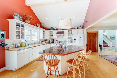 Bright kitchen with white cabinets, a central island, and red accent walls, leading to a staircase and hallway.