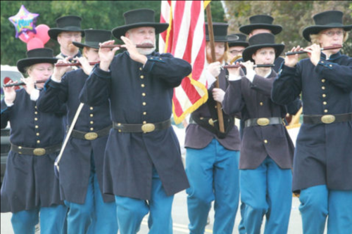 A marching band in historical uniforms plays flutes, with an American flag in the background during a parade.