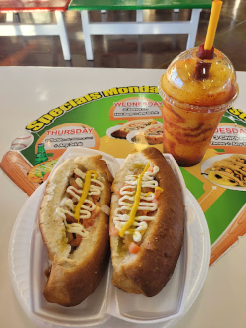 Two hot dogs with mustard and mayonnaise on a white plate, alongside a colorful drink, on a promotional table mat.