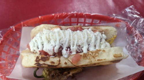 A hot dog in a bun topped with diced tomatoes, mayonnaise, and cheese, served in a red basket.