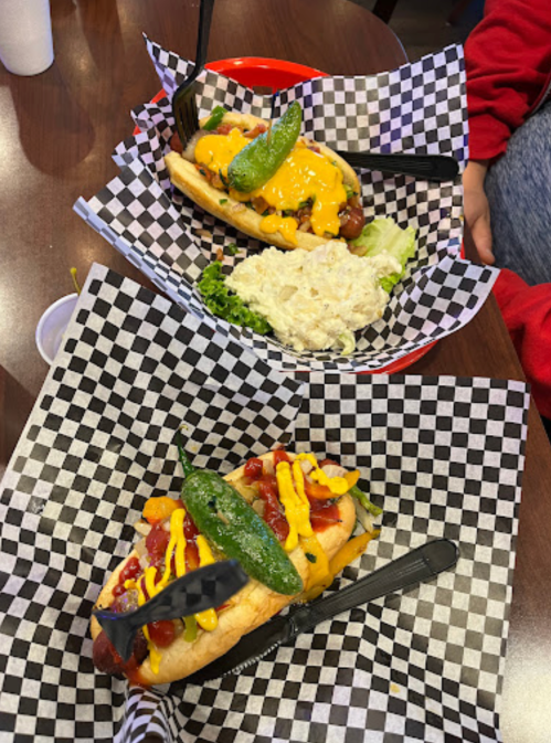 Two loaded hot dogs on checkered paper plates, topped with cheese, jalapeños, and mustard, alongside a side of coleslaw.