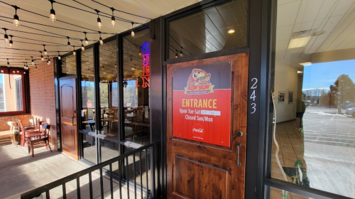 Entrance of a restaurant with a sign indicating hours and an open neon sign, featuring a cozy interior view.