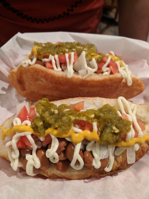 Two loaded hot dogs topped with beans, onions, tomatoes, mustard, mayonnaise, and green sauce on a paper-lined tray.