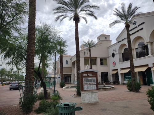 A plaza with palm trees, shops, and a fountain under a cloudy sky.