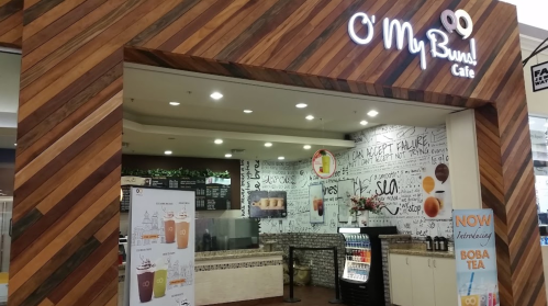 Exterior of O' My Bun! Cafe with wooden decor, menu boards, and a display of drinks, featuring a boba tea promotion.