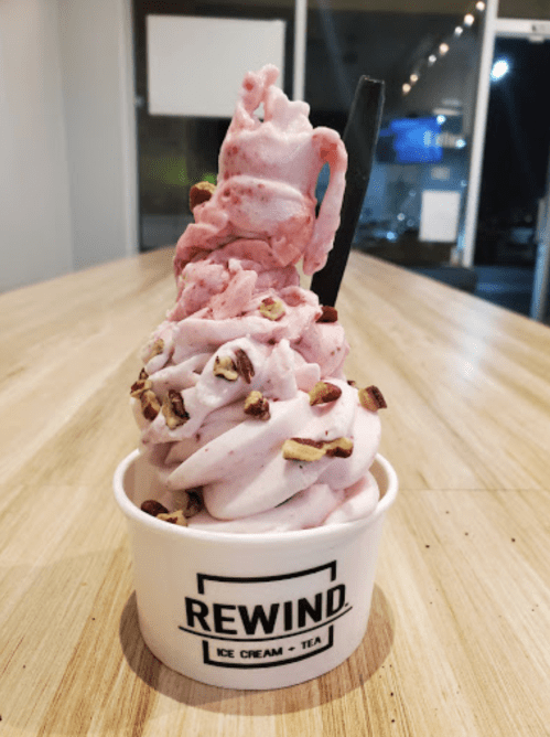 A cup of pink soft serve ice cream topped with chopped nuts, served in a "REWIND" branded container on a wooden table.