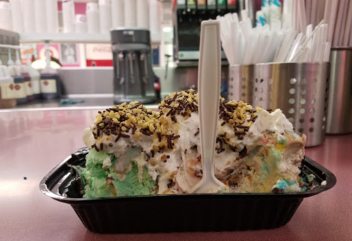 A colorful ice cream sundae topped with whipped cream, sprinkles, and nuts in a black container on a counter.