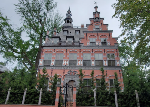 A large, ornate red brick house with a steep roof, decorative details, and surrounded by greenery.